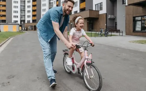 Tata uczy córkę, jak nauczyć dziecko jazdy na rowerze. Radosna dziewczynka na różowym rowerze, tata ją asekuruje.