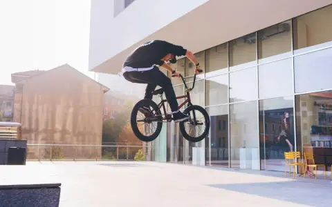Chłopak na BMX-ie wykonuje bunny hop, unosząc rower w powietrze przed nowoczesnym budynkiem.