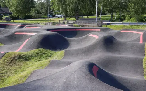 Asfaltowy pump track z czerwonymi pasami, otoczony zielenią i drzewami. Idealne miejsce do zabawy na rowerze lub hulajnodze.