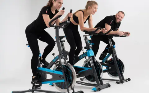 Trzy osoby, dwie kobiety i mężczyzna, ćwiczą na rowerach stacjonarnych. Wszyscy uśmiechnięci, widać, że lubią spinning.