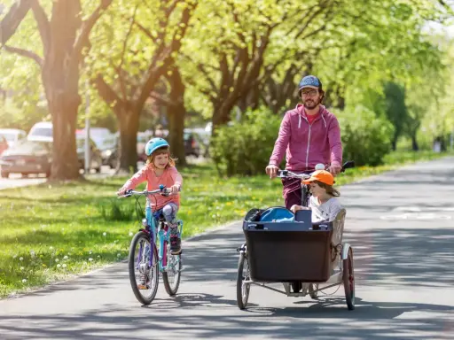 Radosne dziecko na rowerze jedzie przed rodzicem, który wiezie drugie dziecko w przyczepce rowerowej.