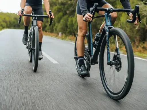 Dwie osoby na rowerach szosowych ROMDO RATT pędzą po asfaltowej drodze wśród zieleni.