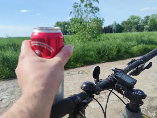 Rowerzysta trzyma puszkę napoju, co sugeruje kierowanie rowerem w stanie nietrzeźwości. Na kierownicy widoczny licznik.