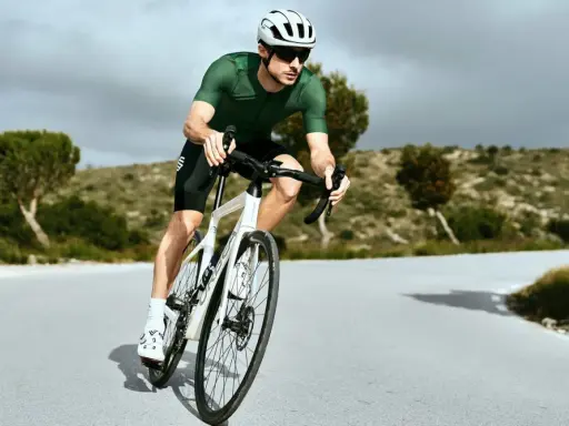 Kolarz w zielonej koszulce i białym kasku w aerodynamicznej pozycji na rowerze szosowym, pokonujący krętą drogę wśród pagórków.