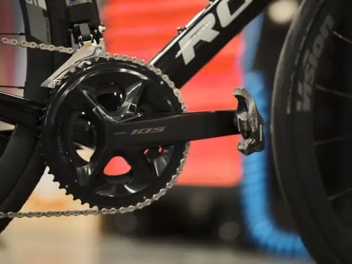 Szukasz informacji, jak dobrać korbę do roweru? Zobacz zbliżenie na czarną korbę Shimano 105 z łańcuchem i pedałem.