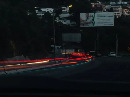 Smugi świateł samochodowych na drodze nocą, sugerujące średnią prędkość. W tle widać domy na wzgórzu i billboard.