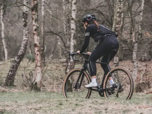 Kobieta w kasku i czarnym stroju jedzie na rowerze typu gravel przez las.