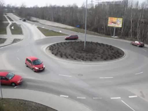 Rowerzysta na rondzie, otoczony przez czerwone samochody i drzewa.