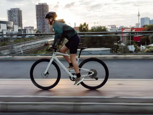 Kobieta w kasku i sportowym stroju jedzie na białym rowerze crossowym po moście, z miejskim krajobrazem w tle.