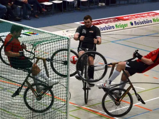 Gracze w piłkę rowerową walczą o piłkę przy bramce. Jeden z zawodników próbuje strzelić gola.