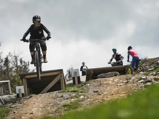 Młoda rowerzystka w kasku i goglach skacze na rowerze w Szczyrk Bike Park. W tle inni rowerzyści.