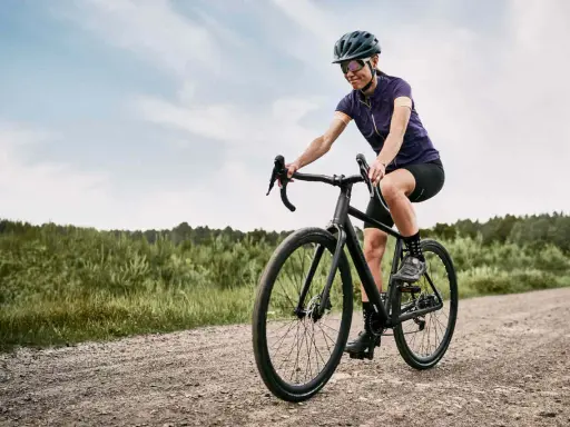 Kobieta na rowerze **gravel** pokonuje szutrową drogę. W tle las i niebo.