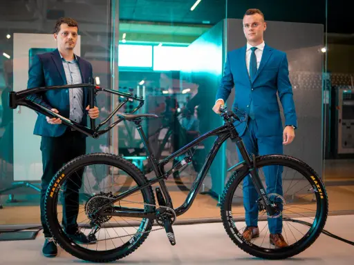Dwóch mężczyzn prezentuje nowy rower full-suspension. Jeden trzyma ramę, drugi stoi przy gotowym rowerze. To duma firmy Romet.