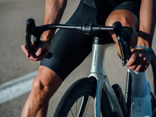 Kolarz w czarnych spodenkach i białych skarpetkach trzyma kierownicę aerodynamicznego roweru Orbea. Szytka rowerowa jest gotowa do jazdy.