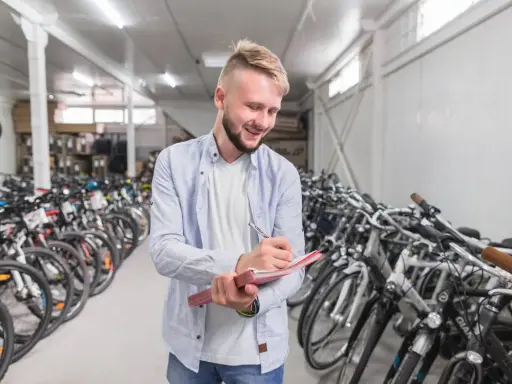 Uśmiechnięty mężczyzna notuje w zeszycie, otoczony rzędami rowerów różnych marek w magazynie.