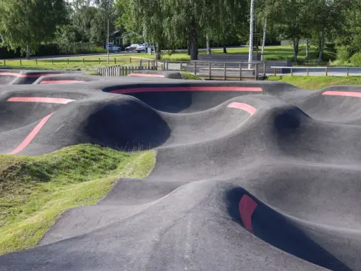 Asfaltowy pump track z czerwonymi pasami, otoczony zielenią i drzewami. Idealne miejsce do zabawy na rowerze lub hulajnodze.