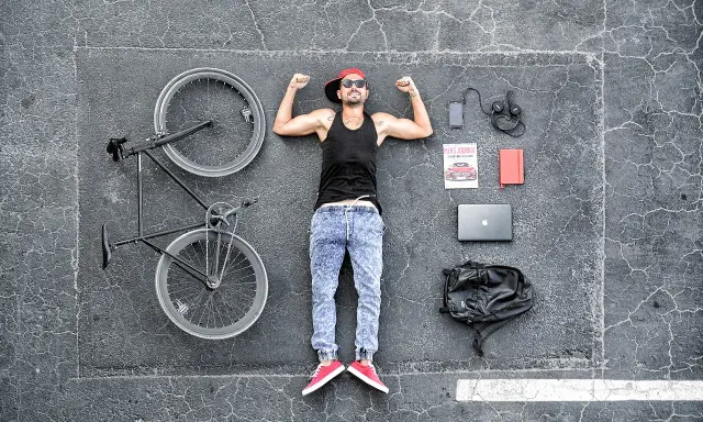 Gotowy na **rowerem do pracy**? Mężczyzna z rowerem, laptopem, telefonem i plecakiem leży na asfalcie.
