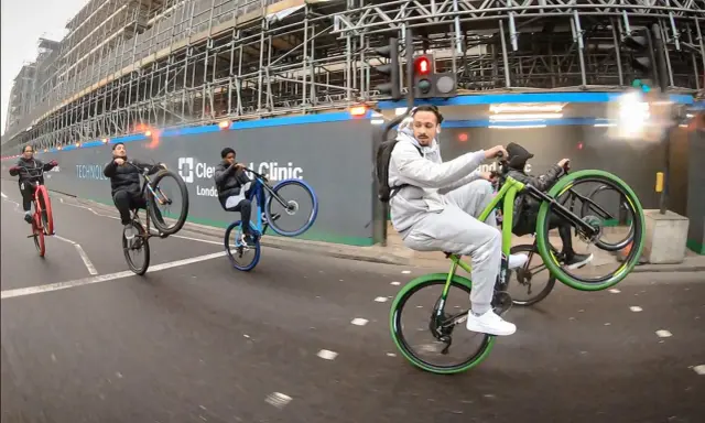 Młodzi mężczyźni wykonują **jazdę na jednym kole** na rowerach, mijając plac budowy. Jeden z nich, w szarym dresie, robi to na zielonym rowerze.