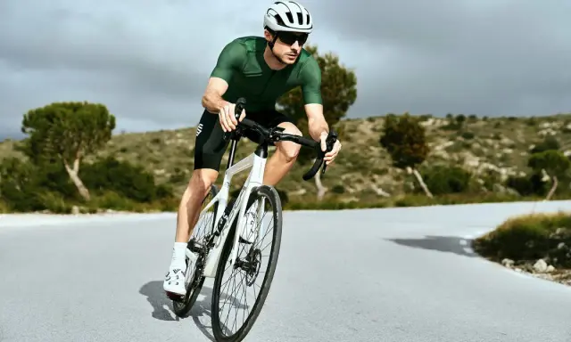 Kolarz w zielonej koszulce i białym kasku w aerodynamicznej pozycji na rowerze szosowym, pokonujący krętą drogę wśród pagórków.