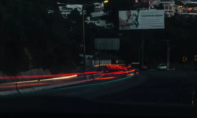 Smugi świateł samochodowych na drodze nocą, sugerujące średnią prędkość. W tle widać domy na wzgórzu i billboard.