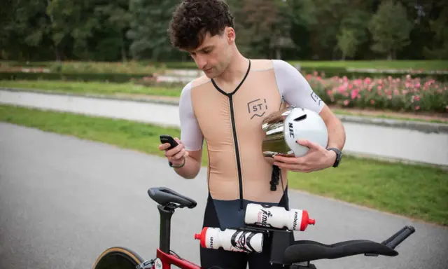 Kolarz sprawdza telefon, trzymając kask. Zastanawia się, co to jest kadencja, gdy patrzy na rower.