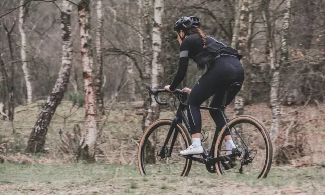 Kobieta w kasku i czarnym stroju jedzie na rowerze typu gravel przez las.
