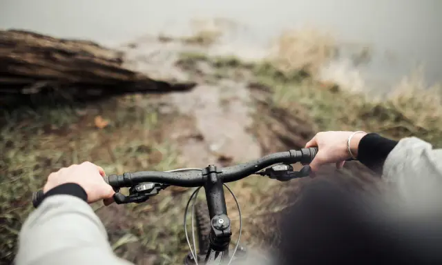 Ręce na kierownicy roweru, błotnista ścieżka i widok na wodę. To właśnie dlatego warto jeździć rowerem – bliskość natury i wolność.