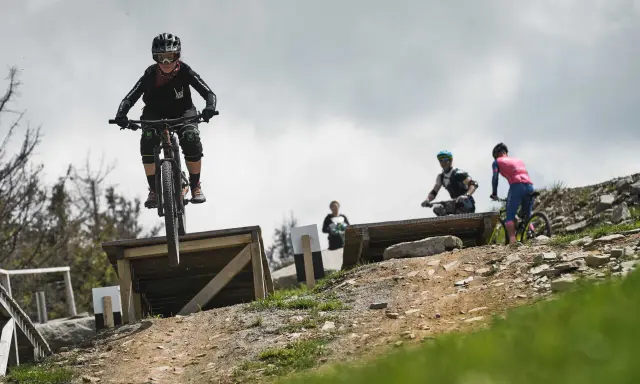 Młoda rowerzystka w kasku i goglach skacze na rowerze w Szczyrk Bike Park. W tle inni rowerzyści.