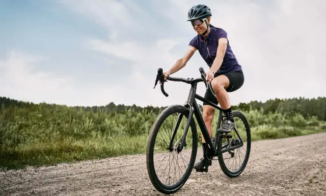 Kobieta na rowerze **gravel** pokonuje szutrową drogę. W tle las i niebo.