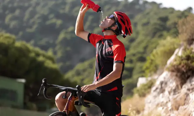 Kolarz w kasku i stroju sportowym pije wodę z bidonu, odpoczywając po intensywnym treningu, który podnosi VO2 max.