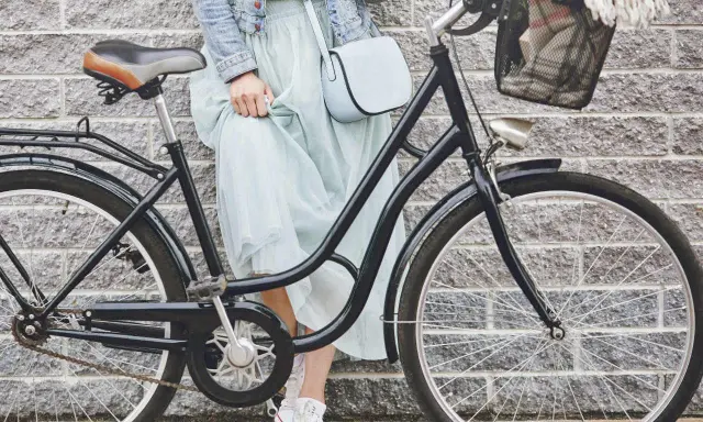 Elegancka kobieta w zwiewnej sukience i dżinsowej kurtce opiera się o rower. Widoczny rozmiar ramy roweru sugeruje, że jest to model miejski, idealny na przejażdżki po mieście.