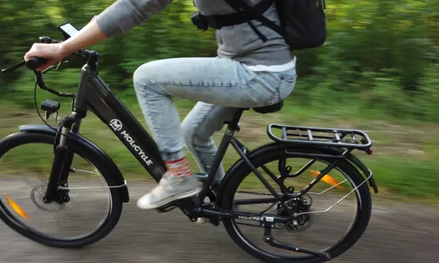 Osoba na rowerze Molicyle, który wygląda jak rower miejski czy trekkingowy, jedzie po ścieżce wśród zieleni.