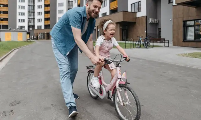 Tata uczy córkę, jak nauczyć dziecko jazdy na rowerze. Radosna dziewczynka na różowym rowerze, tata ją asekuruje.