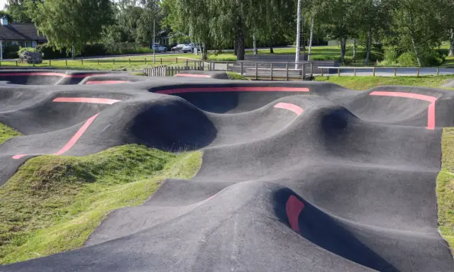 Asfaltowy pump track z czerwonymi pasami, otoczony zielenią i drzewami. Idealne miejsce do zabawy na rowerze lub hulajnodze.