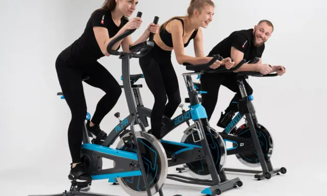 Trzy osoby, dwie kobiety i mężczyzna, ćwiczą na rowerach stacjonarnych. Wszyscy uśmiechnięci, widać, że lubią spinning.