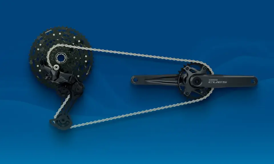 Zestaw napędowy Shimano CUES: korba, przerzutka i kaseta z łańcuchem na niebieskim tle.