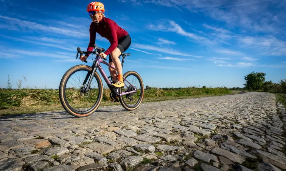 Kobieta w kasku i okularach jedzie na rowerze BMC URS LT po brukowanej drodze.