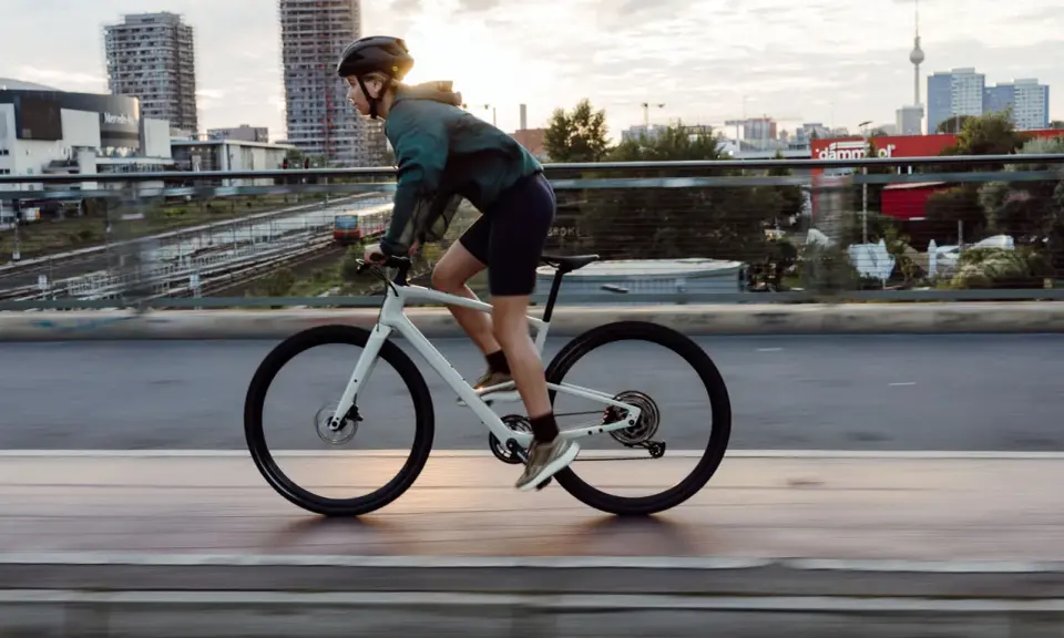 Kobieta w kasku i sportowym stroju jedzie na białym rowerze crossowym po moście, z miejskim krajobrazem w tle.