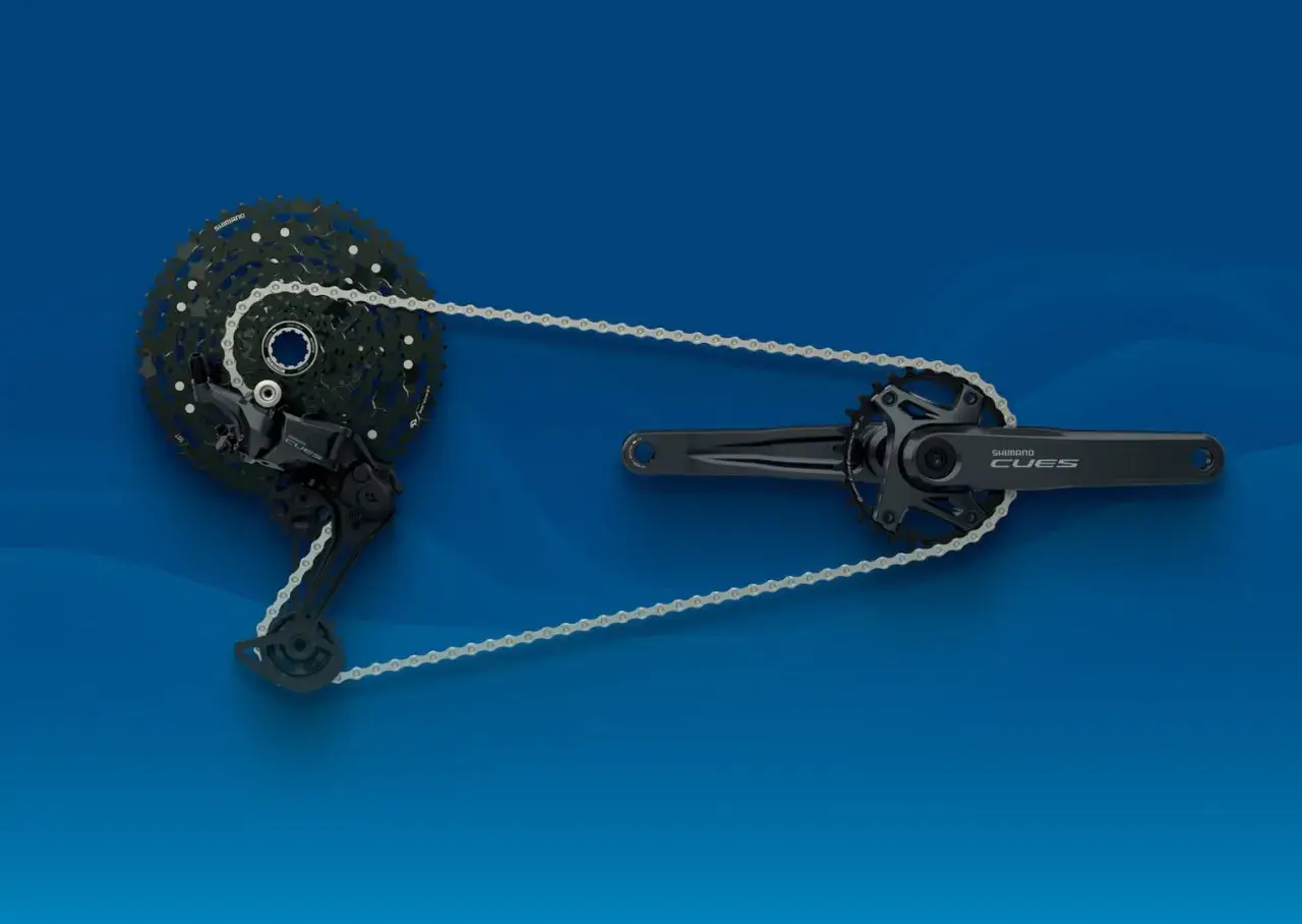 Zestaw napędowy Shimano CUES: korba, przerzutka i kaseta z łańcuchem na niebieskim tle.