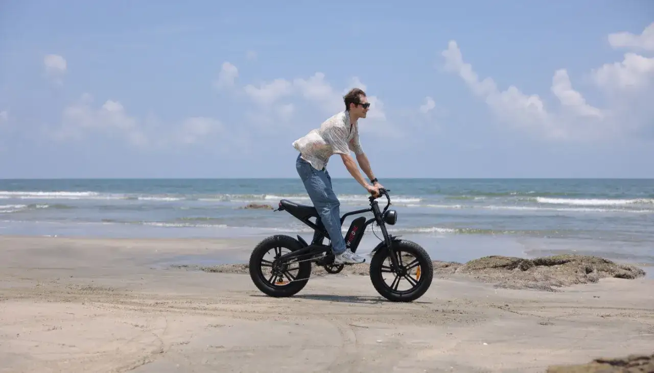 Mężczyzna na plaży, cieszący się jazdą rowerem elektrycznym bez wspomagania. W tle spokojne morze i błękitne niebo.