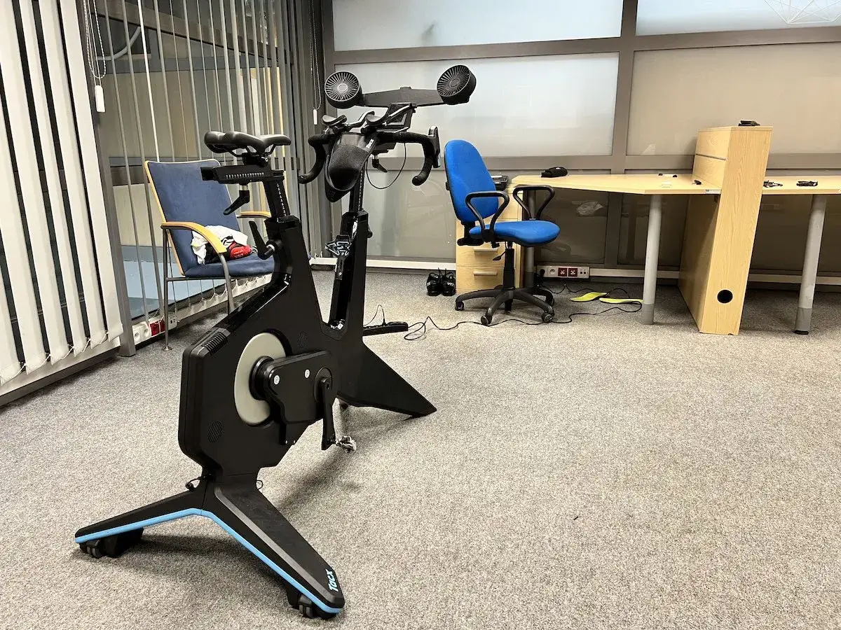 Nowoczesny trenażer rowerowy Tacx, idealny do treningów FTP w domu. Obok biurko i krzesła biurowe.