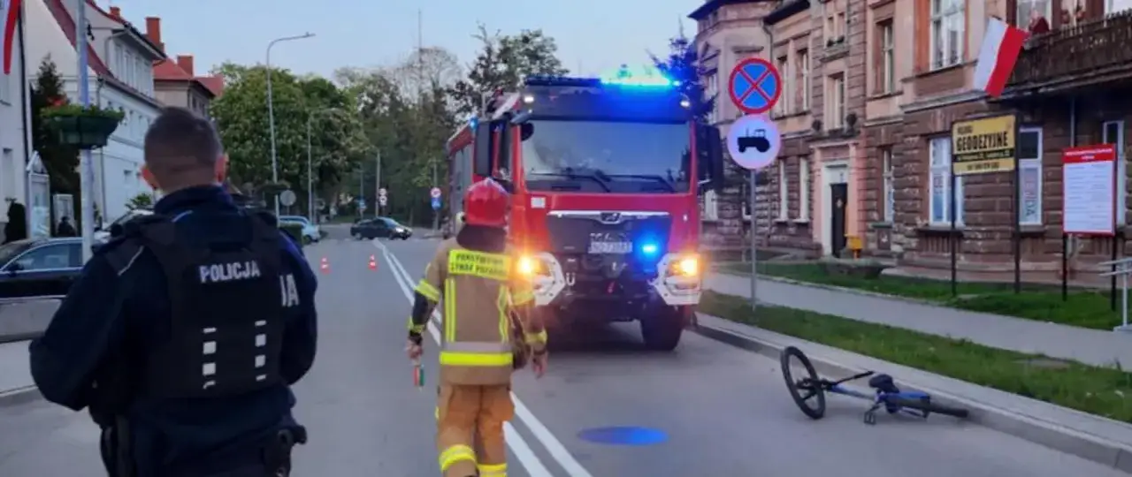 Policjant i strażak przyglądają się leżącemu rowerowi po wypadku. W tle wóz strażacki.