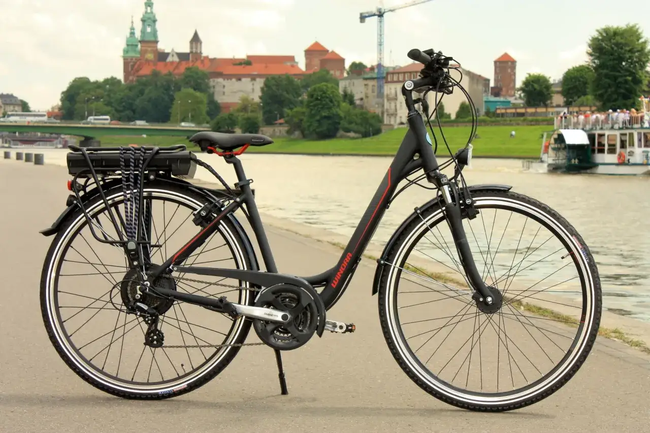 Czarny rower z baterią na bagażniku, gotowy do przeróbki na elektryczny. W tle Wawel i Wisła.
