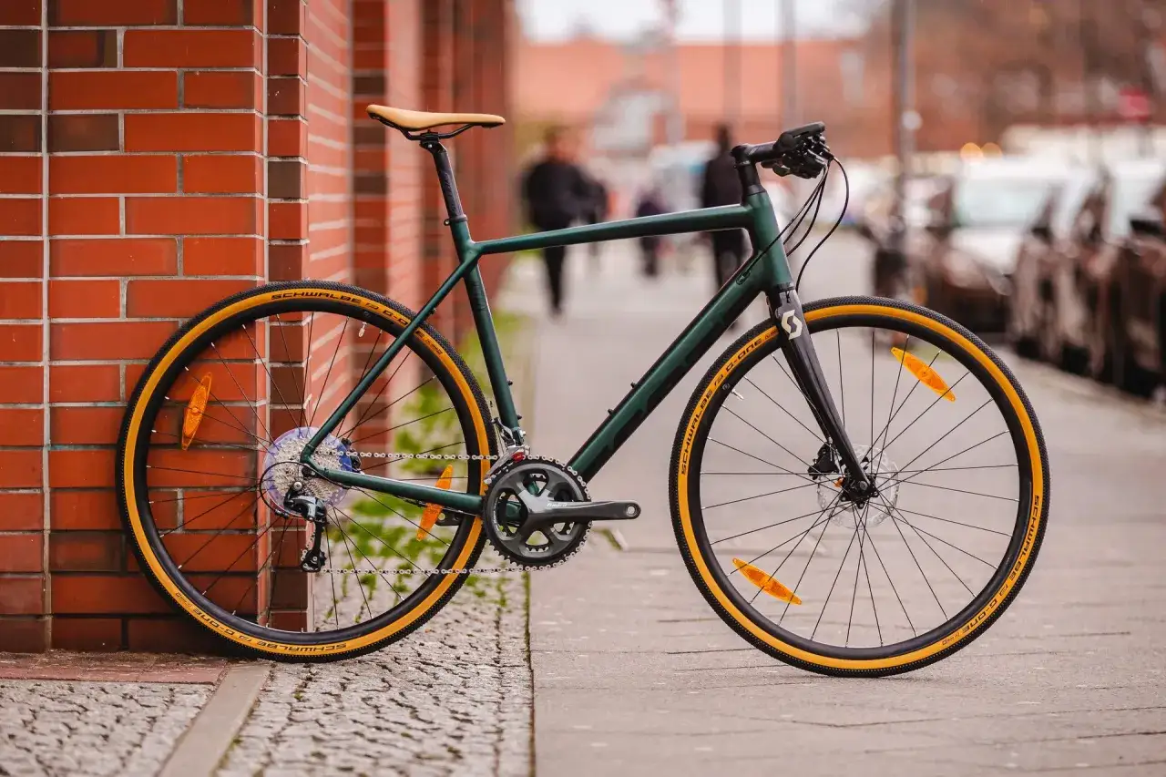 Zielony rower gravel z prostą kierownicą i żółtymi oponami Schwalbe G-One, oparty o ceglaną ścianę.
