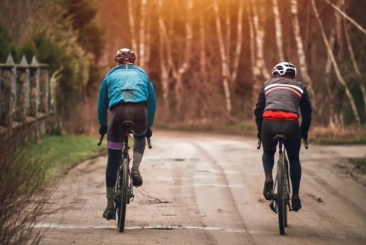 Dwóch rowerzystów w kurtkach i długich spodniach, idealnie jak się ubrać na 15 stopni, jedzie po błotnistej drodze wśród drzew.