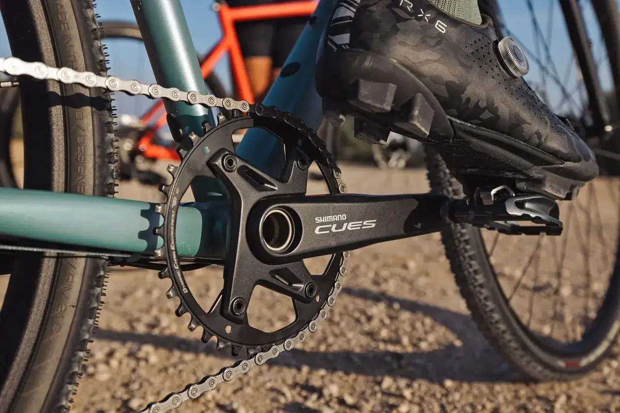 Zbliżenie na rower z napędem Shimano CUES. Widoczna korba, łańcuch i but rowerowy.