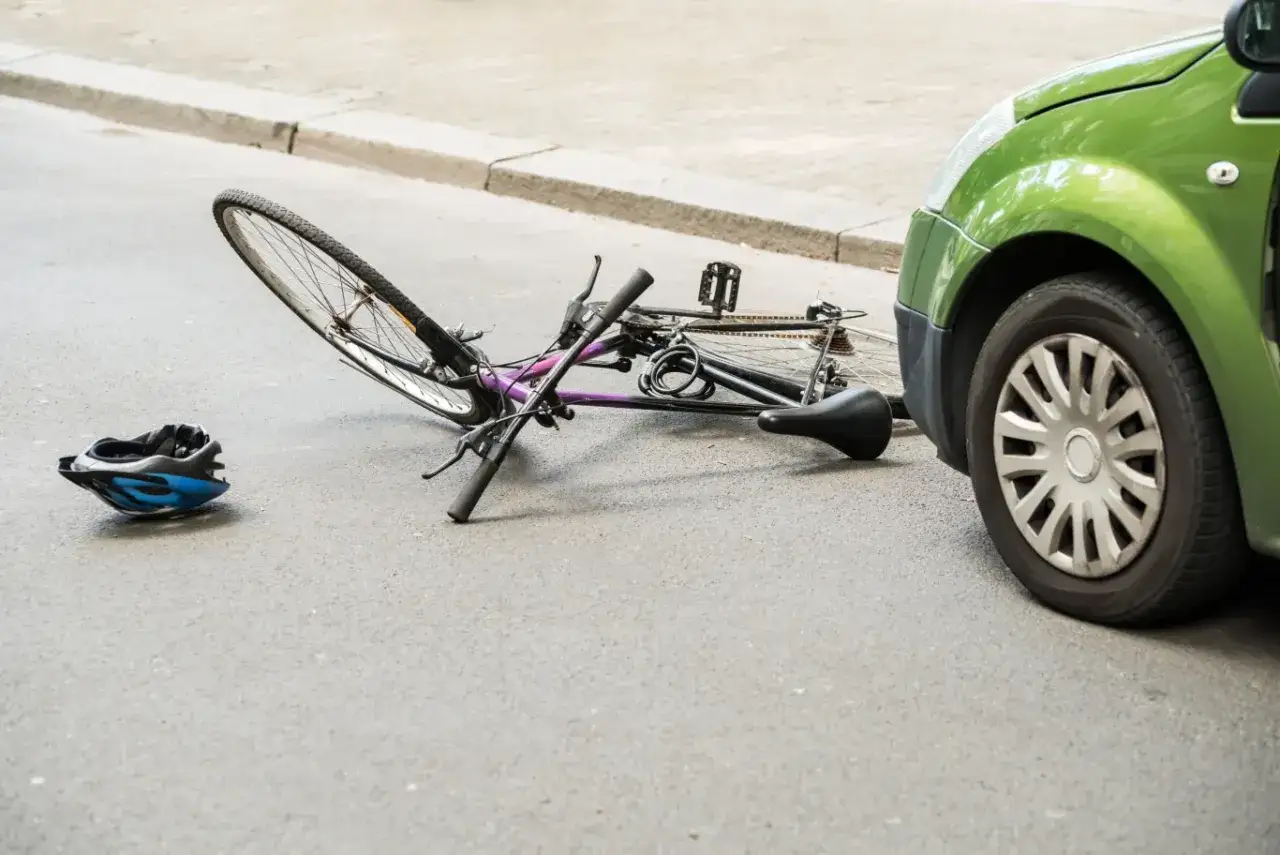 Złamany rower i kask leżą na ulicy obok zielonego samochodu. Wygląda na to, że doszło do wypadku rowerzysty.