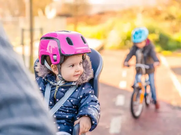 Mała dziewczynka w różowym kasku dla 1,5 rocznego dziecka siedzi w foteliku rowerowym. W tle jedzie inne dziecko na rowerze.
